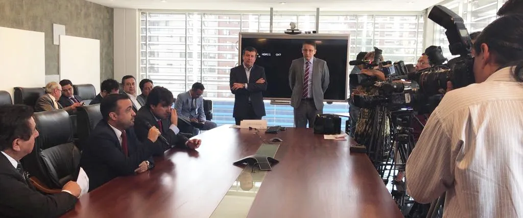 A meeting in a boardroom, with people seated around a long table and television cameras recording.
